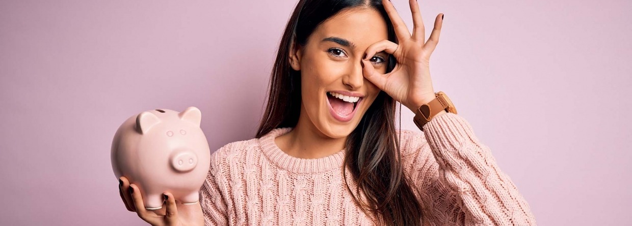 Vrolijke jonge vrouw met een spaarvarken in haar hand en met haar andere hand maakt ze een ok teken voor haar oog staat voor een roze achtergrond