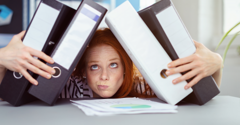 Burn-out; jonge vrouw met haar kin op een stapeltje papieren op haar bureau en houd ordners tegen haar hoofd aan